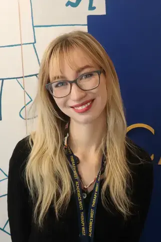 Kiera Mullany, BA, Student Life Coordinator, stands in front of a blue wall, wearing glasses and a black shirt.