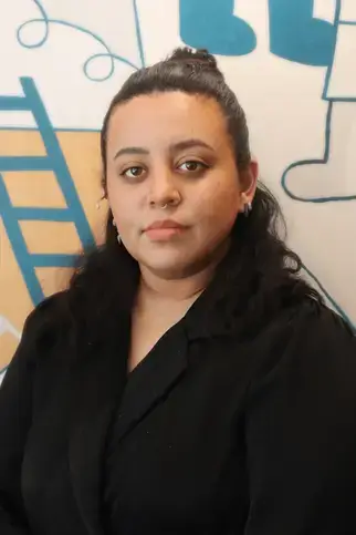 Stephanie Rios, BFA, Upper School Lead Teacher in Art, stands in front of a wall wearing a black shirt and jacket.