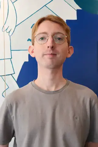 Alex Sirgenson, MA, Head Academic Coordinator, stands in front of a wall, wearing glasses and a striped shirt.